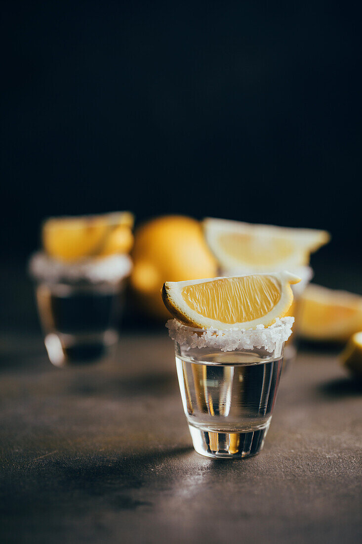 Tequila shots with salt and lemon placed on reflective surface against dark background