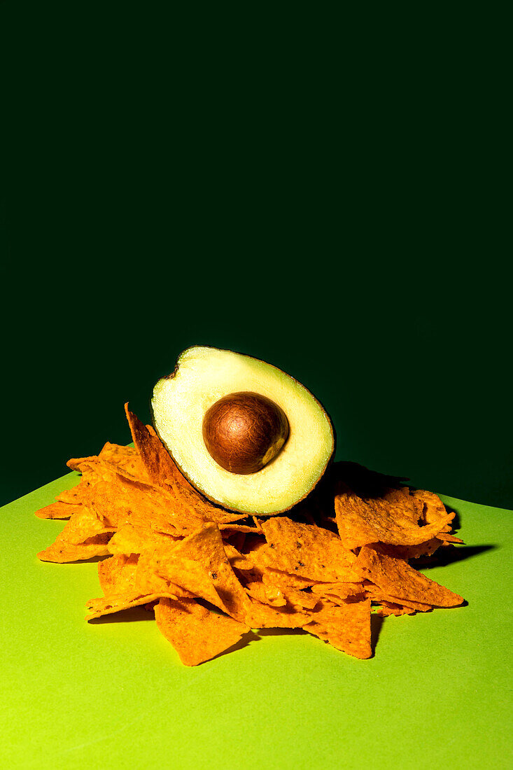 Halbe reife Avocado mit Kern auf einem Stapel knuspriger Tortilla-Chips vor dunkelgrünem Hintergrund