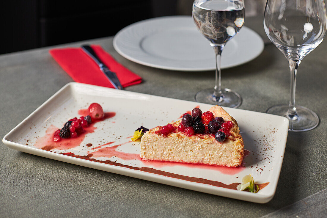 Piece of delicious cheesecake with red berries and jam served on rectangular plate near glasses of water in restaurant