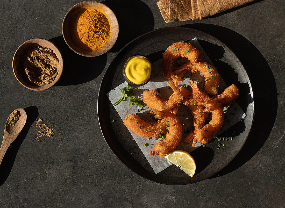 From above still life with plate of lemon and mustard battered prawns with two bowls of turmeric and curry spices