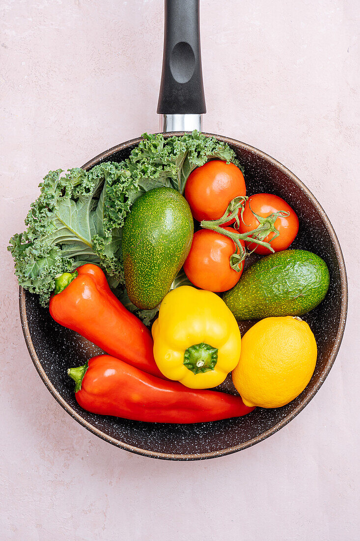 Draufsicht auf Bratpfanne mit rohen reifen Paprika und Avocados neben Kirschtomaten und Grünzeug auf hellem Hintergrund in Küche