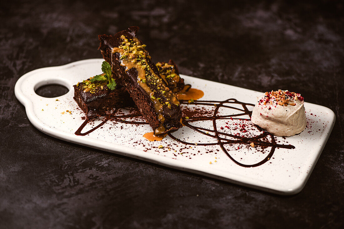 Tasty brownies covered with peanut butter and crunchy crushed pistachios near gelato scoop on plate with chocolate sauce