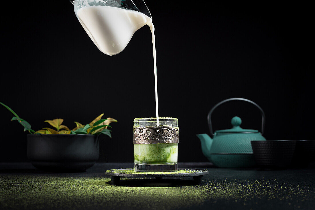 Frische Milch wird aus einem Glas in eine Glastasse mit Matcha-Tee gegossen, die während einer traditionellen Zeremonie auf einem Tisch mit Teekanne und Geschirr steht