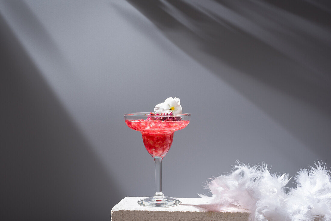 Crystal glass of pomegranate margarita cocktail served with flower blooms near near feathers on concrete blocks in studio