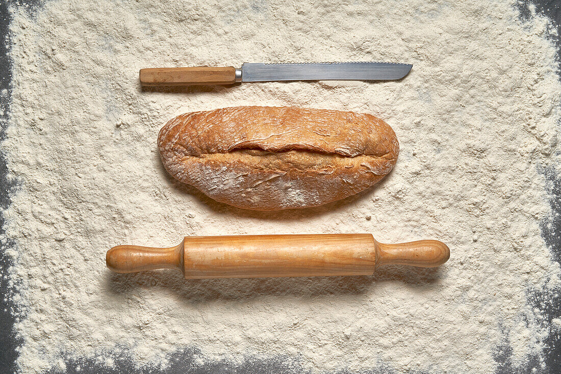 Top view full frame loaf of freshly baked bread with wooden rolling pin and knife placed on white flour in light room