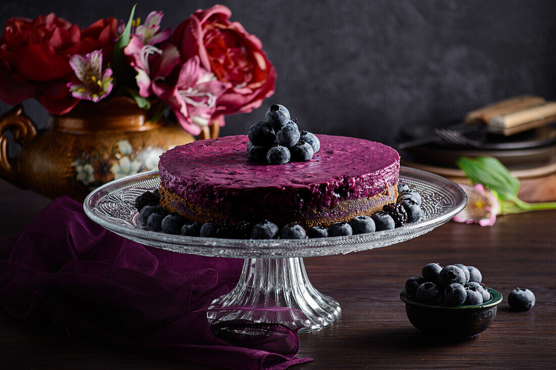 Köstliche Blaubeer-Mousse-Torte mit violetter Sahne, dekoriert mit frischen Beeren, serviert auf einem Glasständer auf einem dunklen Tisch