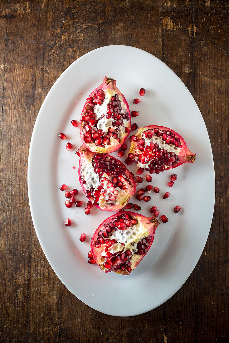Draufsicht auf eine Stilllebenkomposition mit geschnittenen Stücken frischer reifer Granatapfelfrüchte mit Kernen auf einem weißen Teller auf einem Holztisch