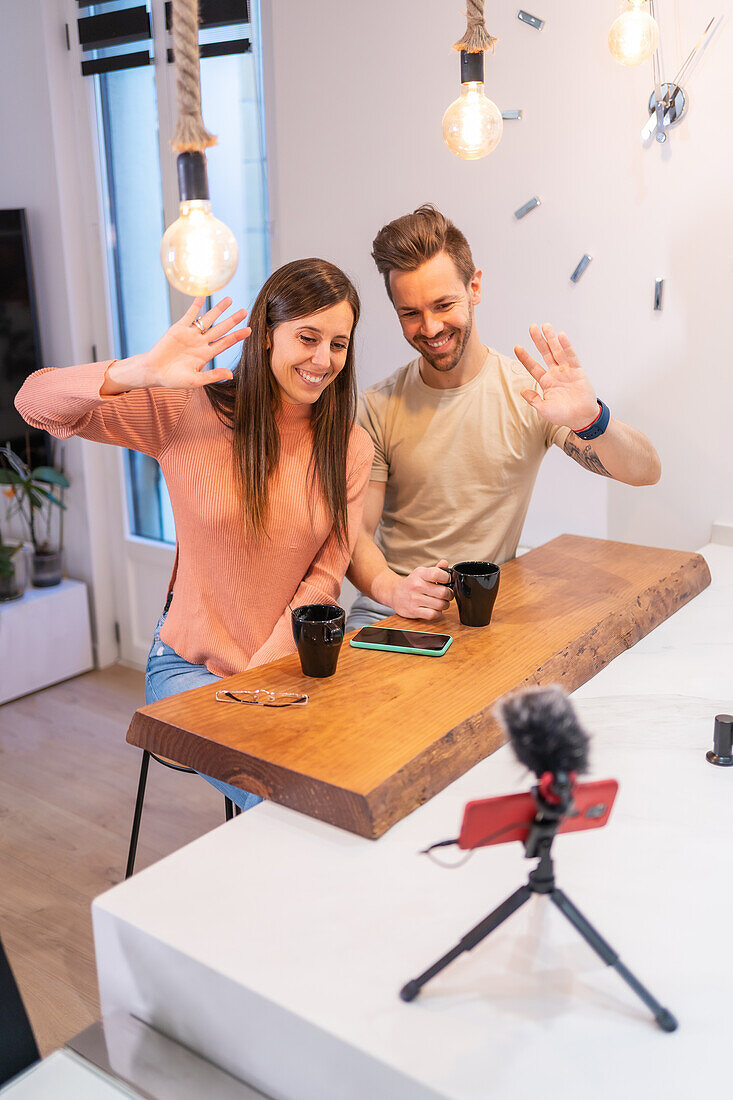 Positive couple of bloggers waving hand to cellphone with microphone while sitting at kitchen counter with cup of coffee at home