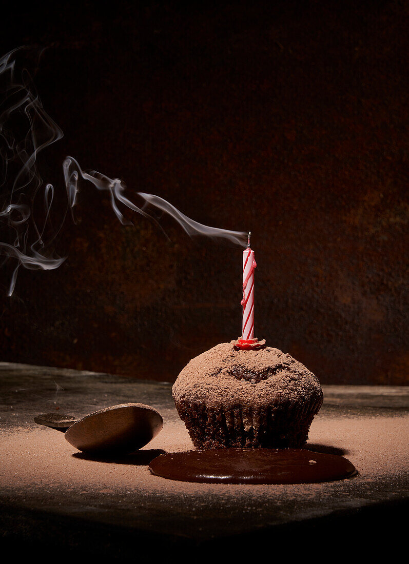 Sweet birthday muffin with powdered sugar and extinguished candle served on table with spilled chocolate and spoon on dark background