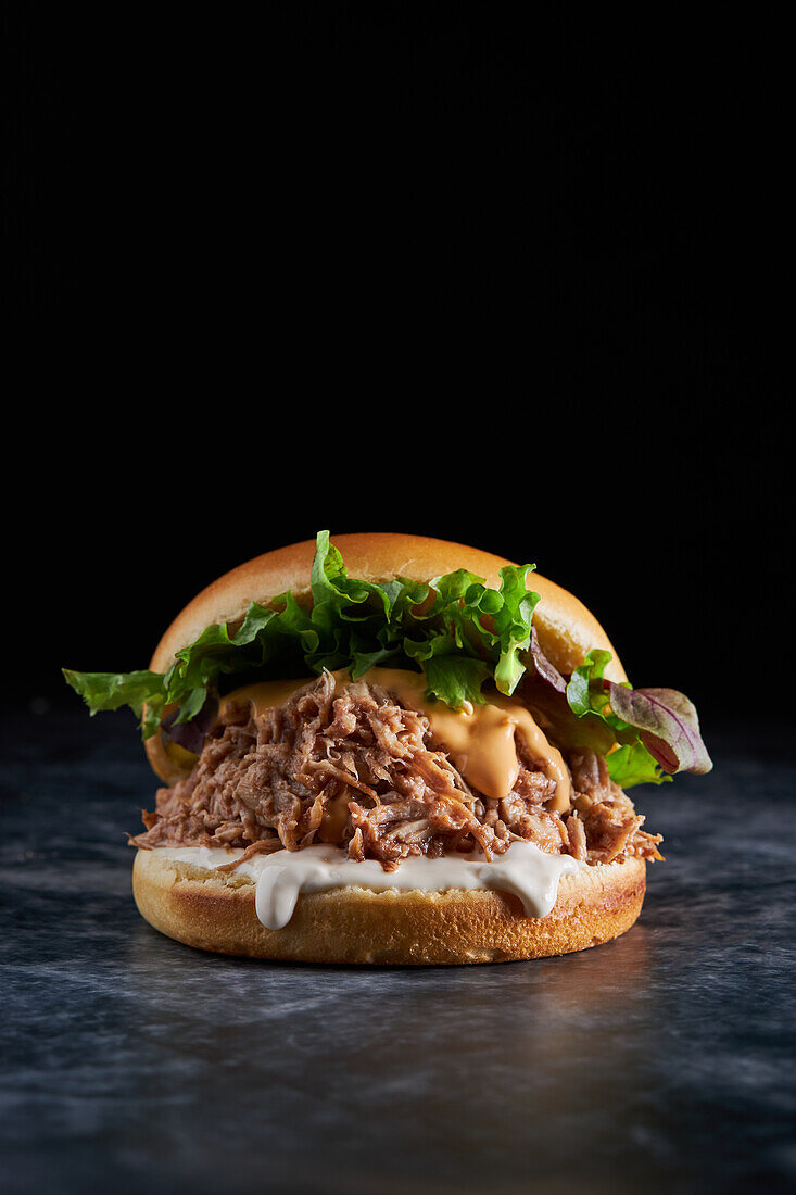 Appetitlicher Burger mit frischem Salat, Käse, Mayonnaise und Schweinefleisch auf einem Tisch in einem dunklen Raum