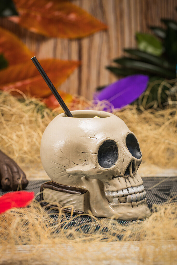 Keramische polynesische Tiki-Tasse in Form eines Totenkopfes mit Strohhalm inmitten von trockenem Gras mit Holzzaun und bunten Federn auf unscharfem Hintergrund