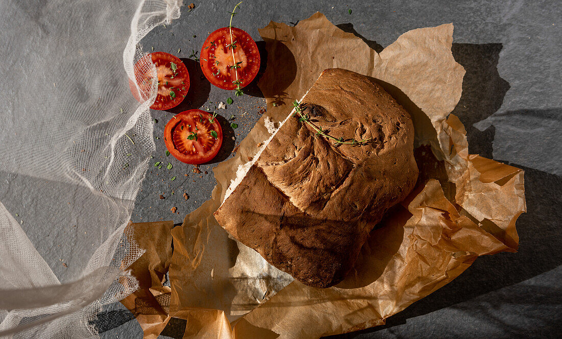 From above of fresh aromatic artisan bread loaf on parchment paper placed near sliced ripe red tomatoes and herbs