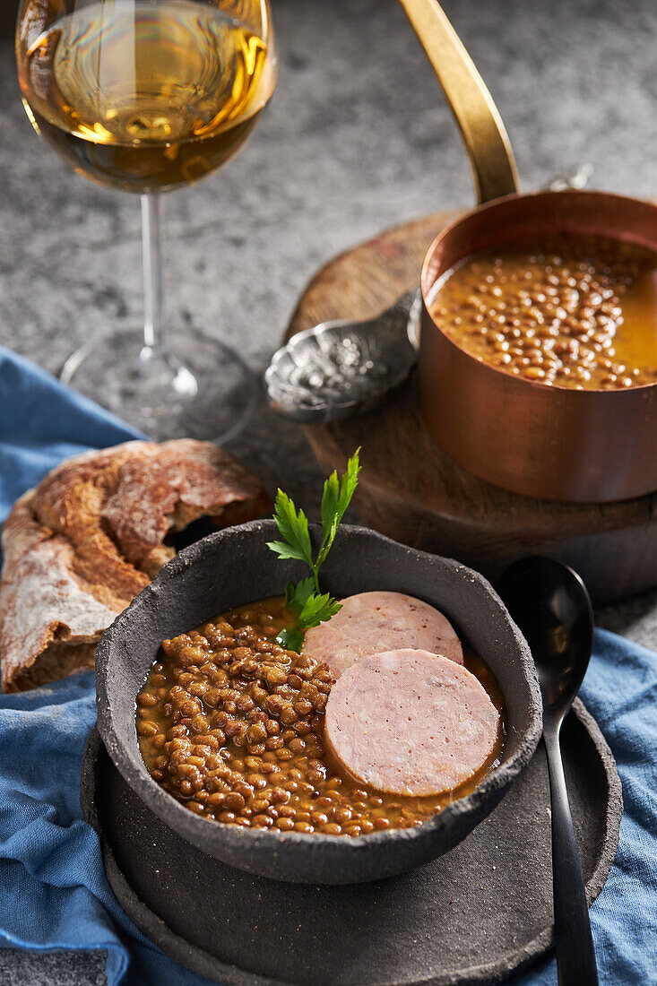 Rustikale Schalen mit leckerer Linsensuppe mit Petersilie und Wurstscheiben auf Marmortisch und blauer Serviette neben Brot und Wein beim Mittagessen