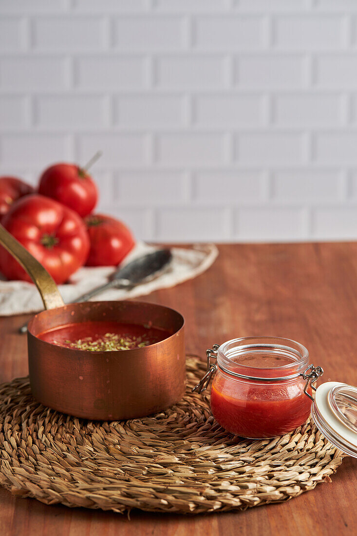 Pan and container with delicious homemade tomato sauce placed on woven mat on wooden table in kitchen