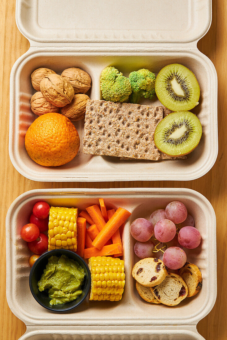 From above of lunch boxes with healthy food including crackers carrot sticks grapes cherry tomatoes with kiwi broccoli walnut and tangerine on yellow background