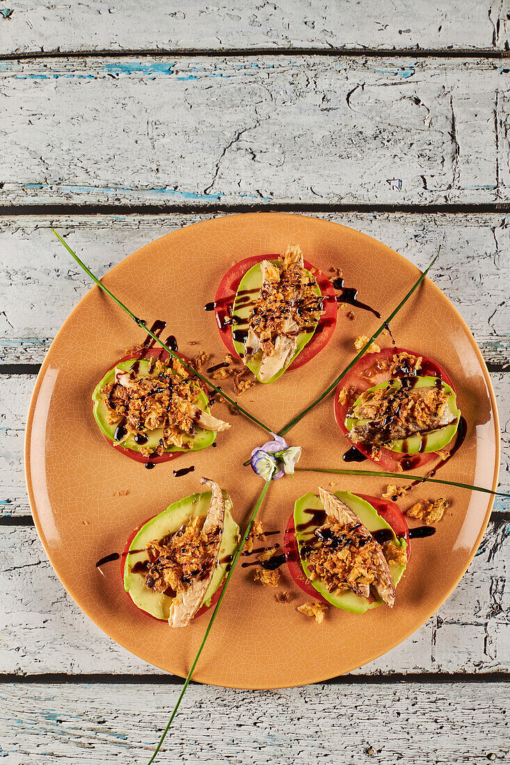 From above crispy fried onion and sliced of mackerel placed on slices of tomato and avocado near greens and flowers on plate in restaurant