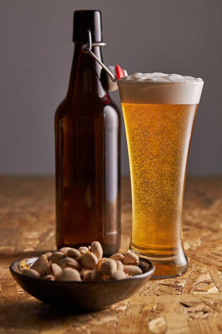 Glas frisches kaltes Bier auf Holztisch neben Flasche und Teller mit Pistazien platziert