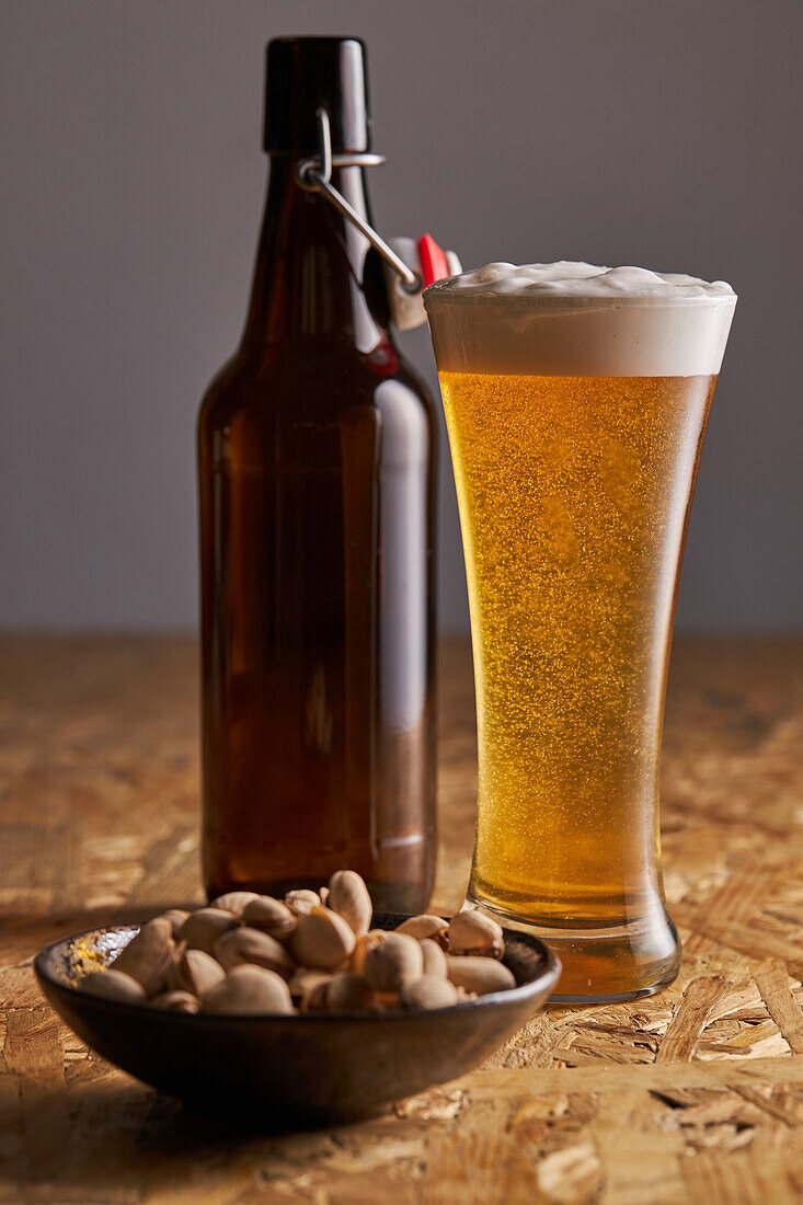 Glass of fresh cold beer placed on wooden table near bottle and plate with pistachios