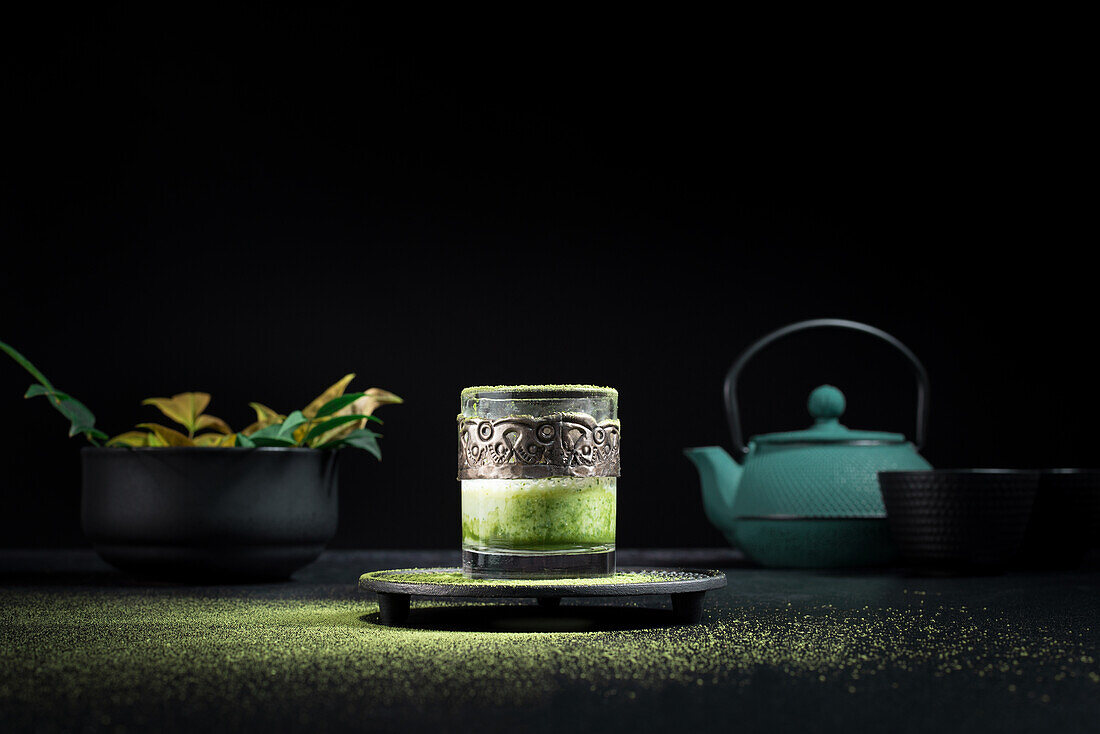 Healthy herbal green matcha tea served in glass cup with metal decoration on saucer sprinkled with powder on black table