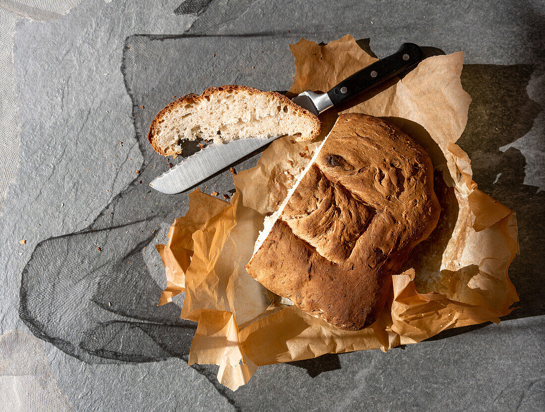 Top view of delicious freshly baked sourdough bread loaf with sliced piece and knife placed on baking paper on gray background