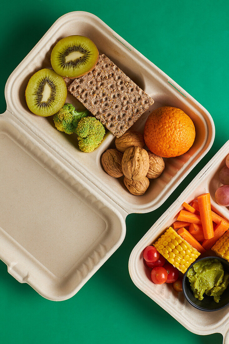 Von oben von Lunch-Boxen mit gesunden Lebensmitteln einschließlich Cracker Karotten-Sticks Trauben Kirschtomaten mit Kiwi Brokkoli Walnuss und Mandarine auf rosa Hintergrund