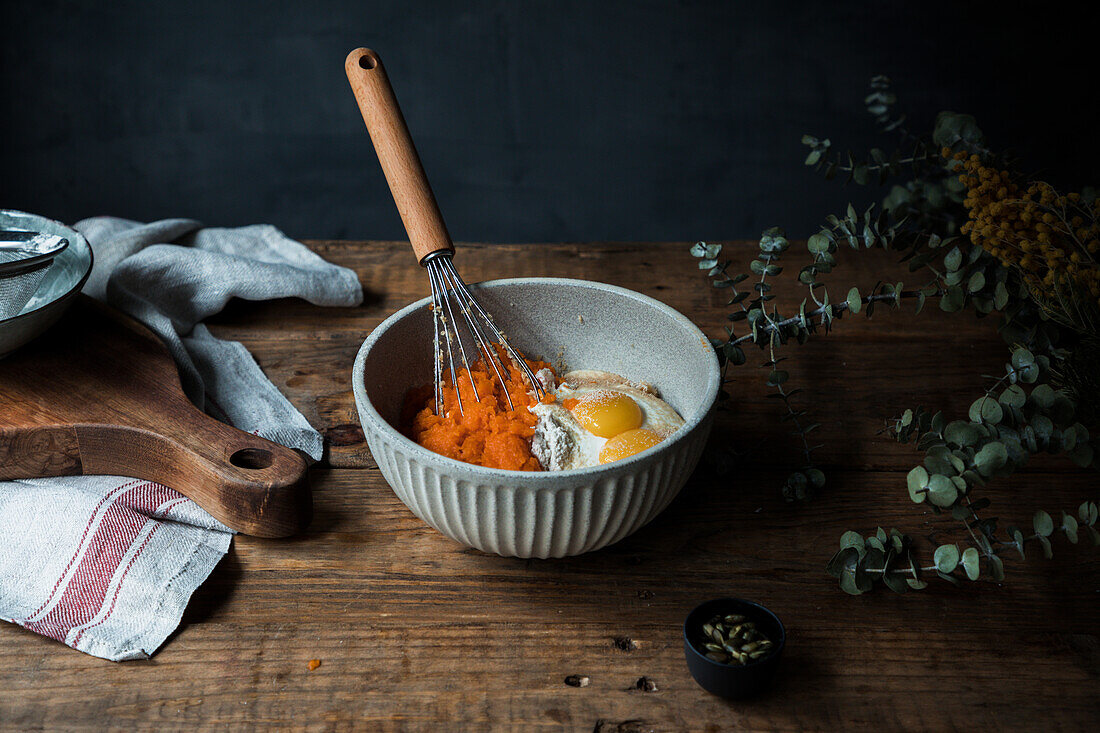 Whisk utensil in a bowl with pumpkin puree, eggs and flour for pie preparation on timber table near cutting board and towel