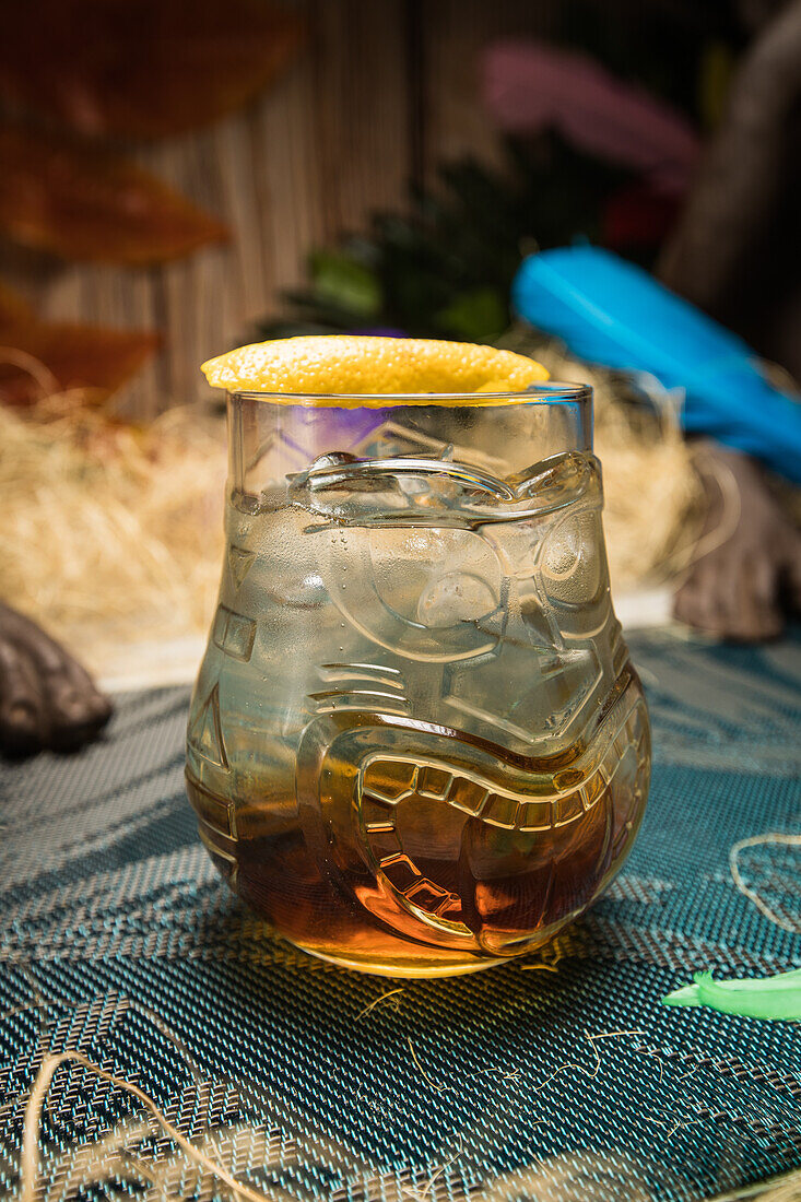 Tiki glass mug with booze placed on edge of wooden table in room with dry grass on blurred background