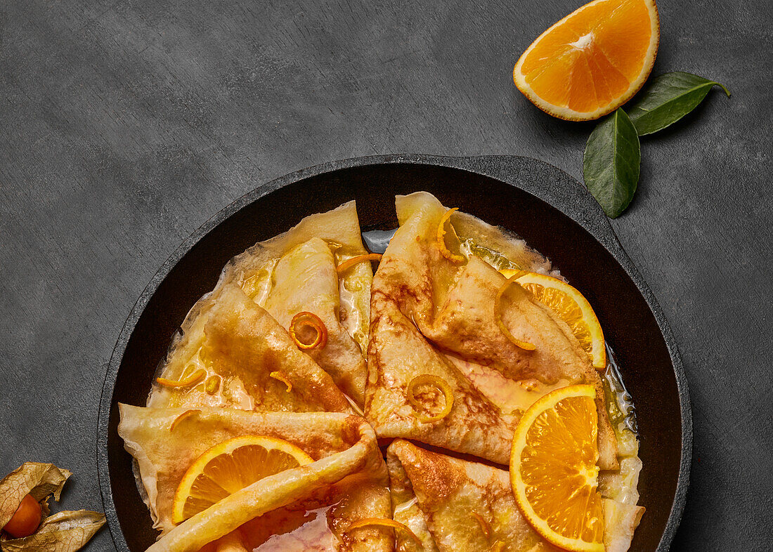 From above homemade crepes suzette with orange liqueur and orange slices served on plate with knife and fork on concrete surface