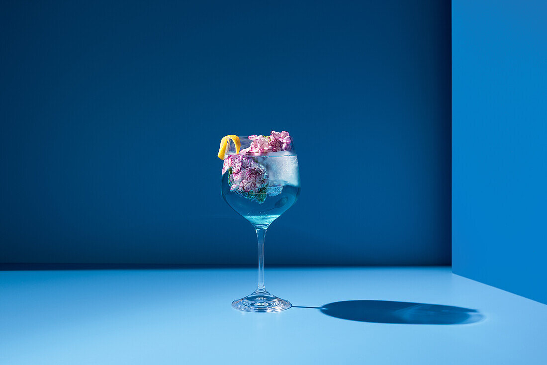 Transparent glass of fresh cocktail with mint leaves and flowers placed on surface against blue background