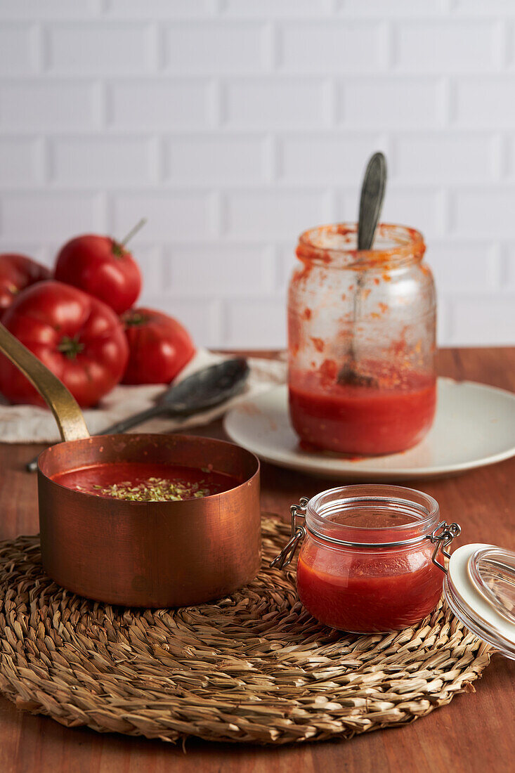 Pfanne und Behälter mit köstlicher hausgemachter Tomatensauce auf einer gewebten Matte auf einem Holztisch in der Küche
