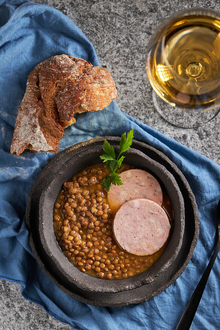 Rustikale Schale mit leckerer Linsensuppe mit Petersilie und Wurstscheiben auf Marmortisch und blauer Serviette neben Brot und Wein beim Mittagessen