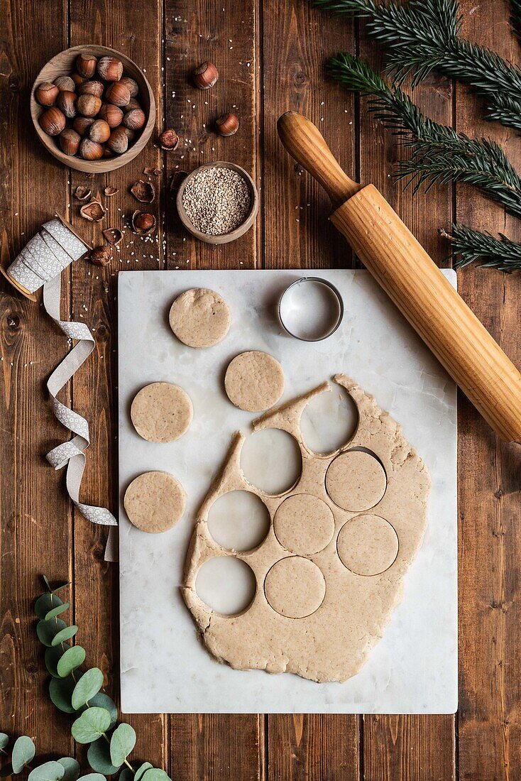 Top view of fresh raw pastry and round shaped cookie cutter placed on table with hazelnuts and Christmas decorations