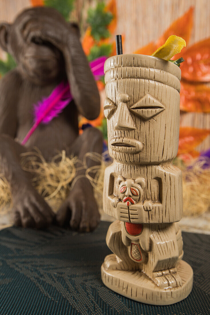 Traditional sculptural tiki cup of alcohol drink with straw placed on rug against wooden fence colorful leaves and dry grass