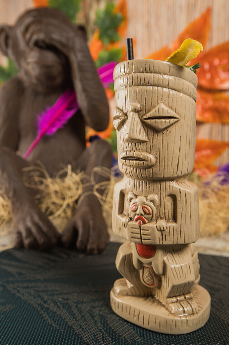 Traditioneller skulpturaler Tiki-Becher mit alkoholischem Getränk und Strohhalm auf Teppich vor Holzzaun mit bunten Blättern und trockenem Gras