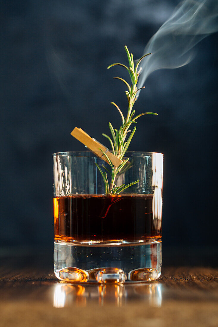 Glass of whiskey with rosemary placed on wooden table against blue background