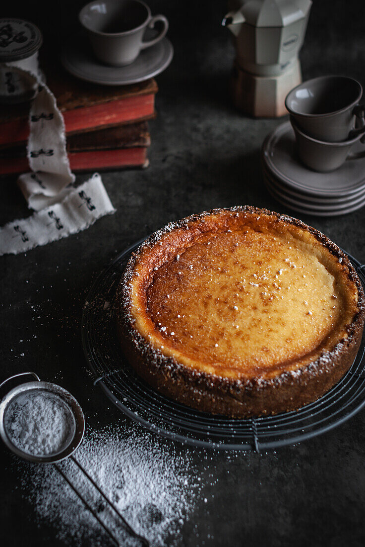 Blick von oben auf einen leckeren hausgemachten aromatischen Käsekuchen mit Puderzucker auf einer Backform neben Kochgeschirr und einem Sieb auf einem schwarzen Tisch