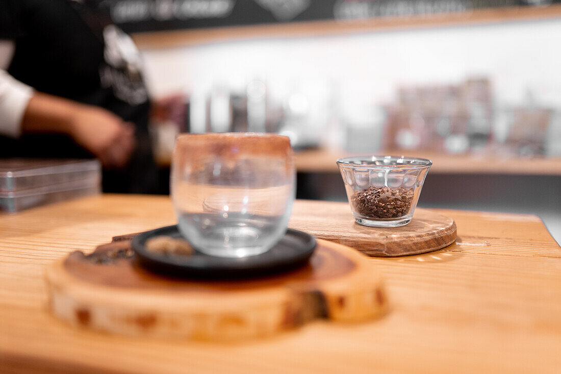 Weichzeichner eines Glases mit zerkleinerten Kakaobohnen auf einem Holzbrett neben einer leeren Tasse auf einem Kaffeetisch
