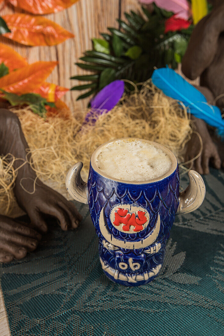 From above of bull shaped tiki mug of alcohol drink with froth placed against dry grass and feathers on blurred background