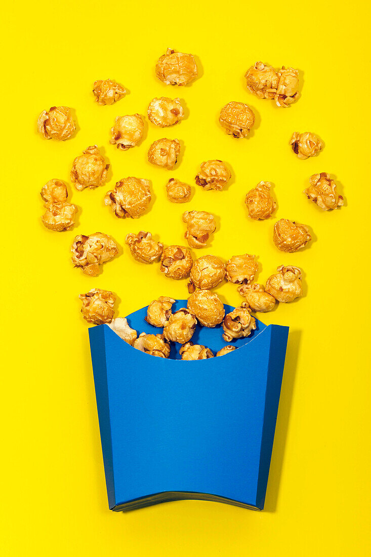 Top view of bright blue paper bag with delicious scattered sweet caramel popcorn on yellow background