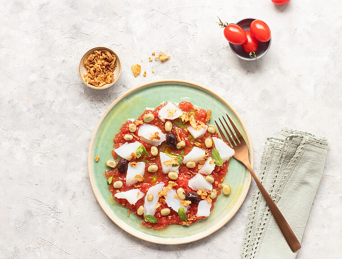 Sommerlicher Tomaten- und Kabeljausalat in einer flachen Schale mit einer Gabel und einer Schüssel mit Röstzwiebeln