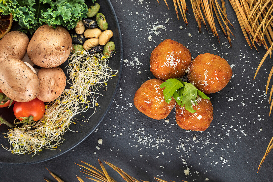 Top view of tasty deep fried croquettes with garlic powder and fresh herb leaves near plate with assorted vegetables
