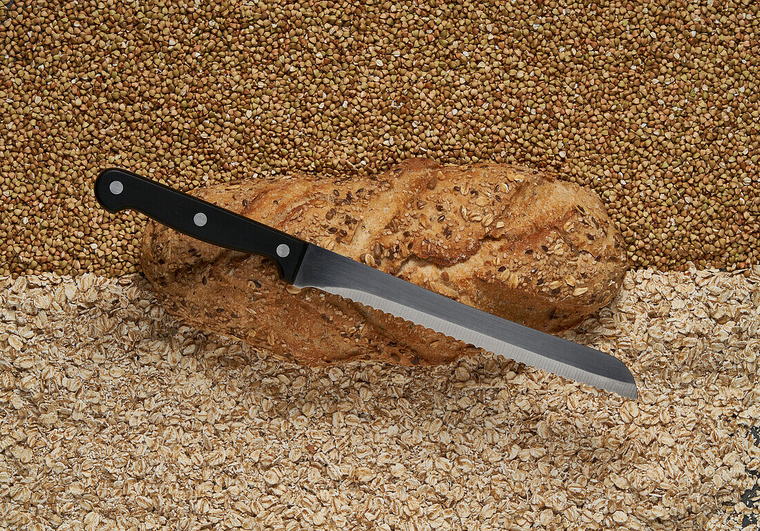 Full frame top view of freshly baked buckwheat and oatmeal bread with knife on top placed on raw grains in light room