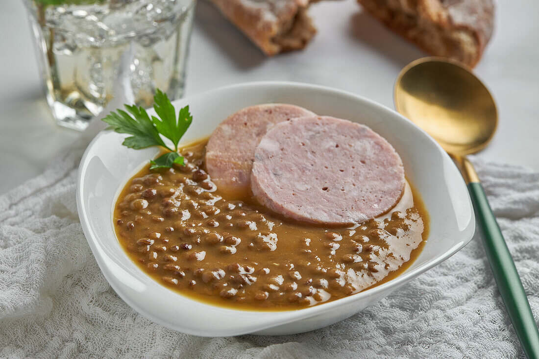 From above spoon placed near bowl of yummy lentil soup served with slices of sausage and leaf of parsley on marble table and gray napkin