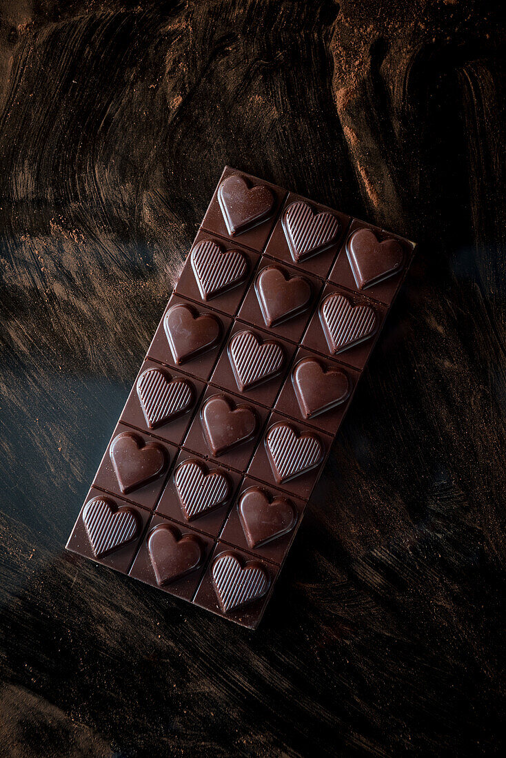 From above of whole chocolate bar with heart shaped decoration served on black background with scattered cocoa powder
