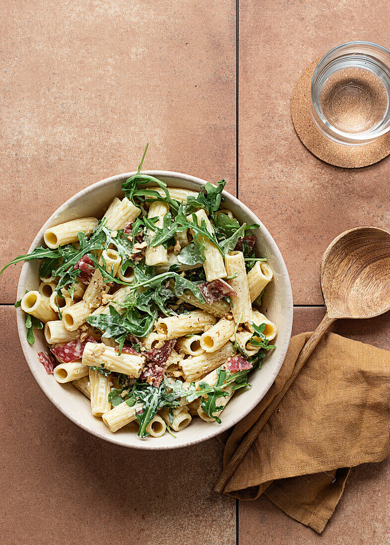 Nudelgericht mit Rucola, Salami und Walnüssen von oben auf einem Tisch, umgeben von Öl, Zitrone und Besteck