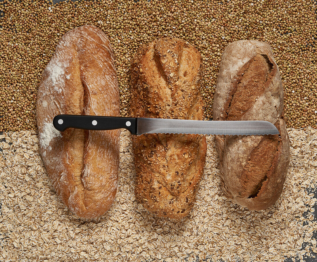 Draufsicht auf köstliche, frisch gebackene braune Brote mit Messer auf rohen Buchweizen und Haferkörnern in heller Küche