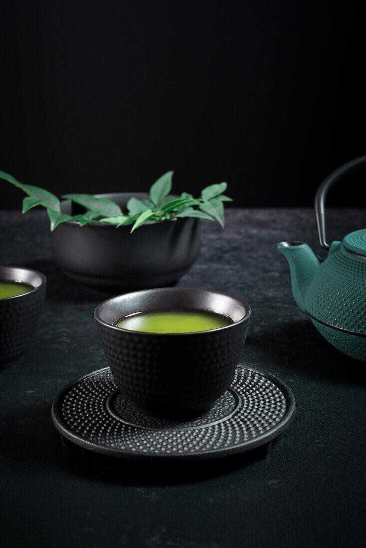 Schwarze Keramiktasse mit traditionellem japanischen grün gefärbten Matcha-Tee, serviert auf einem Tisch mit Teekanne