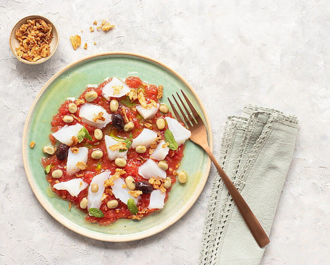 Sommerlicher Tomaten- und Kabeljausalat in einer flachen Schale mit einer Gabel und einer Schüssel mit Röstzwiebeln