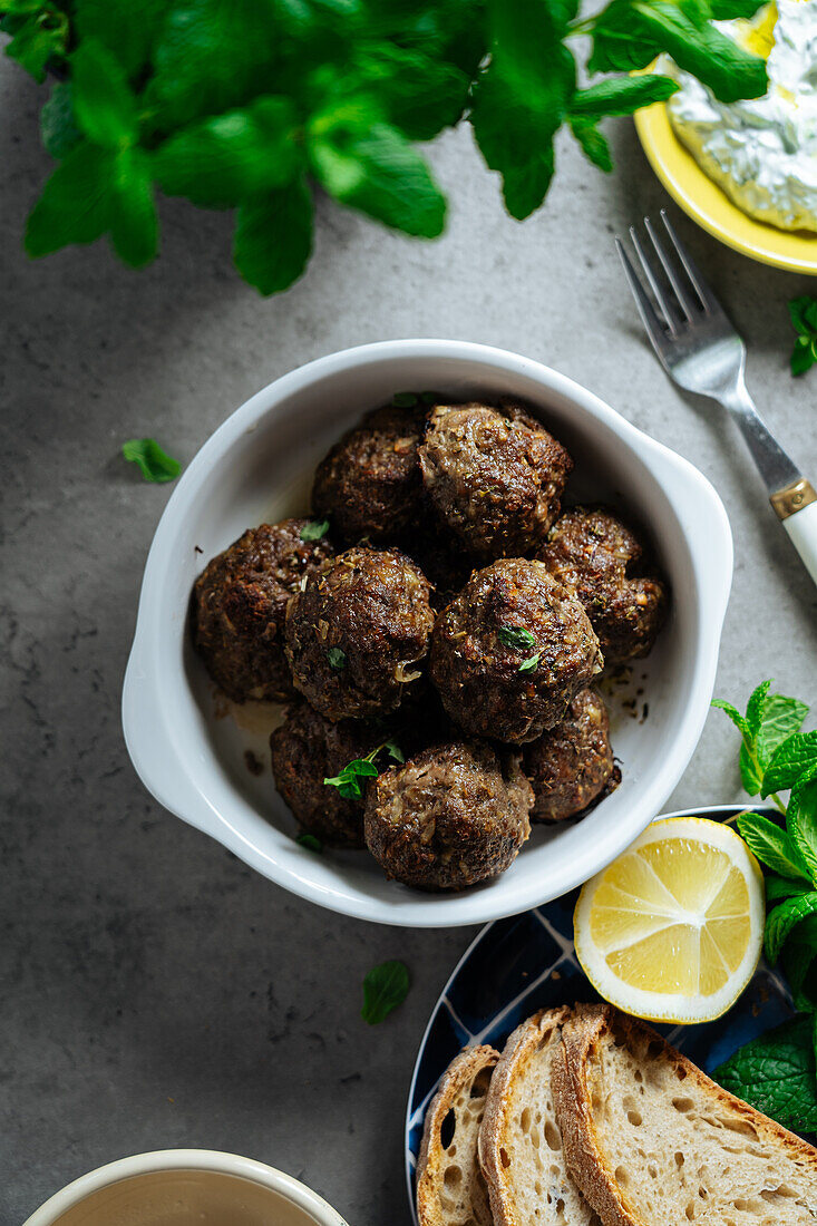 Draufsicht auf appetitliche traditionelle hausgemachte gebratene griechische Fleischbällchen, die auf grauem Hintergrund neben einem Teller mit Brot und Zitrone in der Küche serviert werden