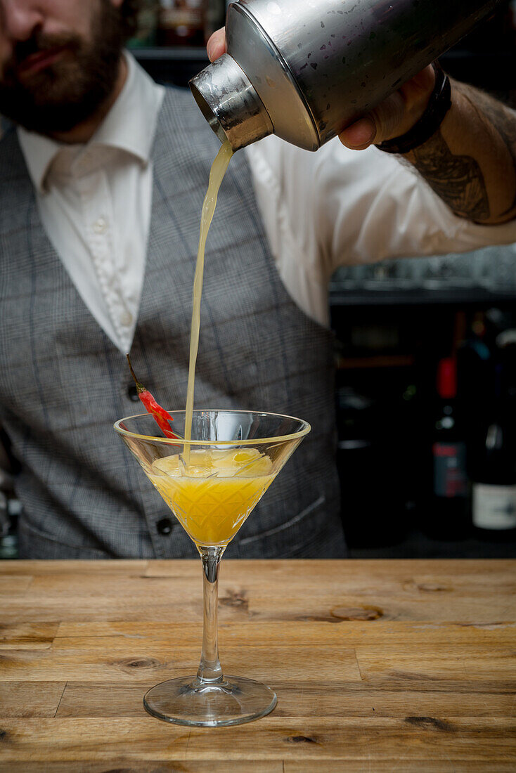 Crop unrecognizable barman pouring alcohol orange cocktail from shaker in glass placed on wooden counter in bar